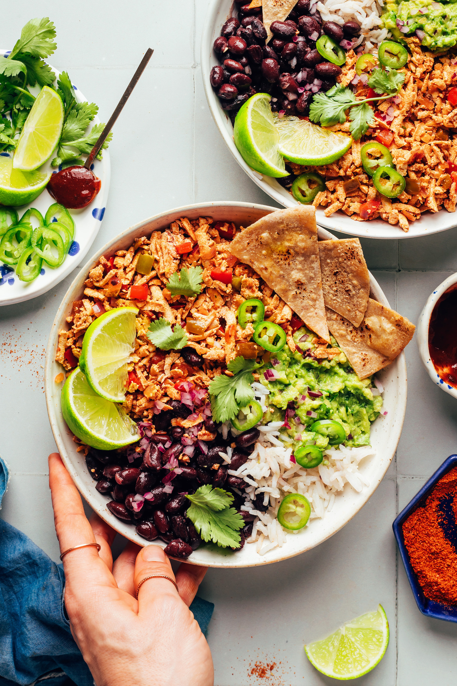 Hand placing down a bowl with rice, black beans, limes, tofu taco meat, tortilla chips, jalapeños, and guacamole