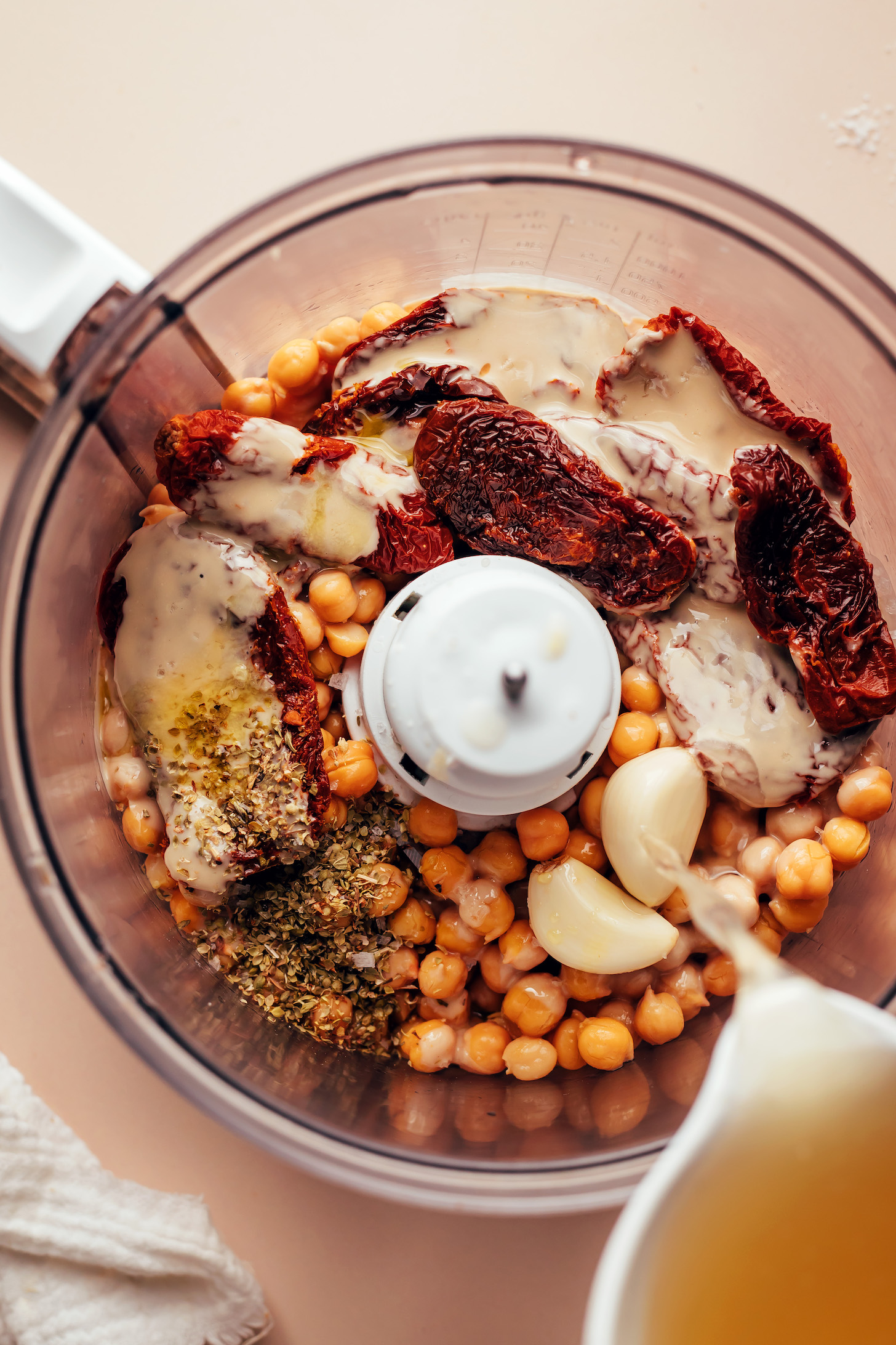 Pouring chickpea brine into a food processor over garlic, chickpeas, sun-dried tomatoes, tahini, olive oil, and dried oregano