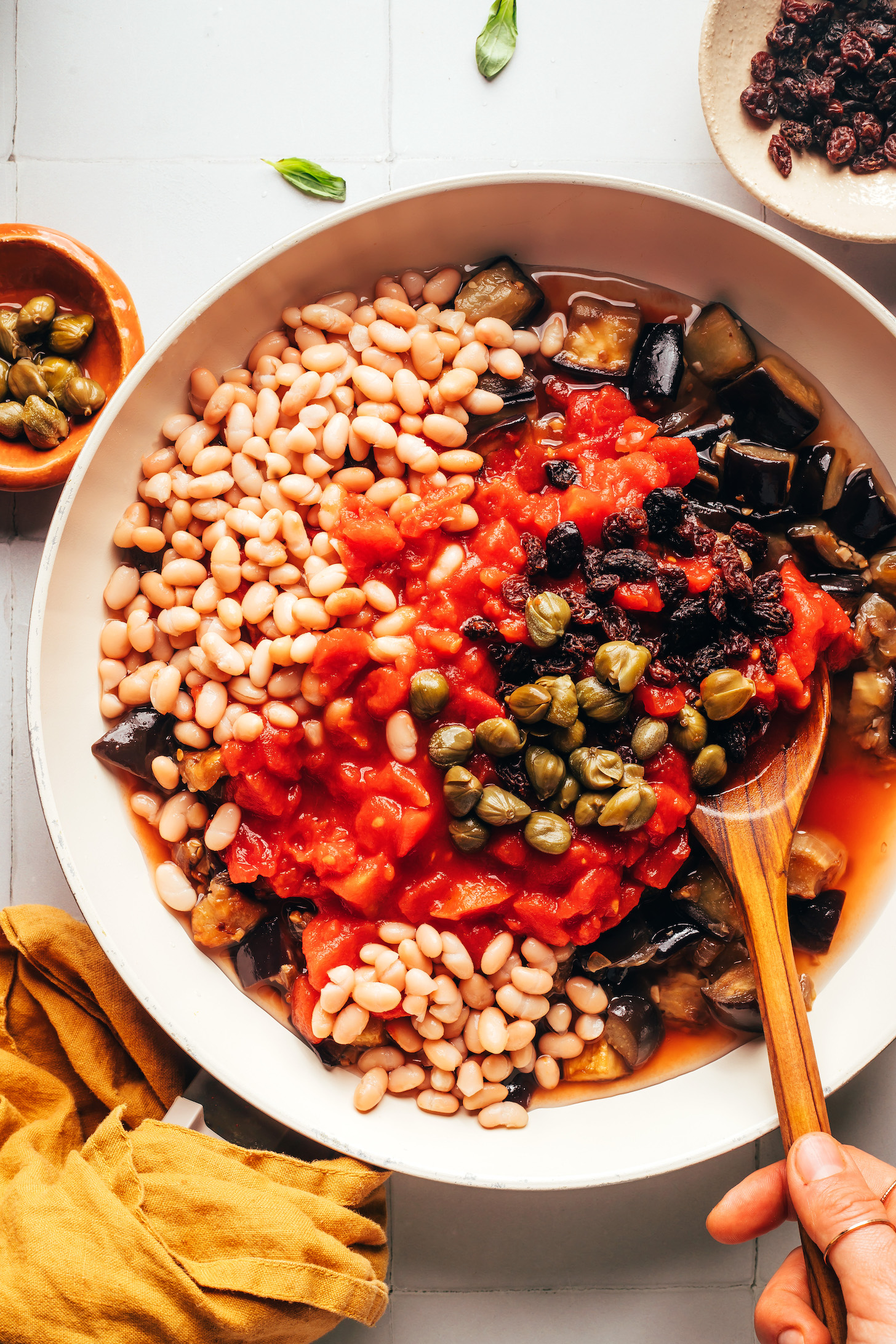 Stirring a skillet of sautéed eggplant, crushed tomatoes, raisins, white beans, and capers